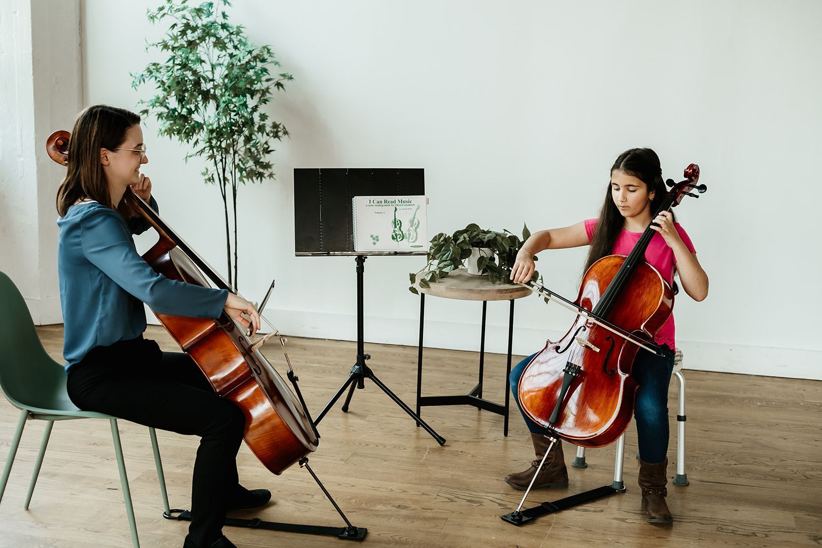 A young boy is playing a cello with his teacher in front of a fence