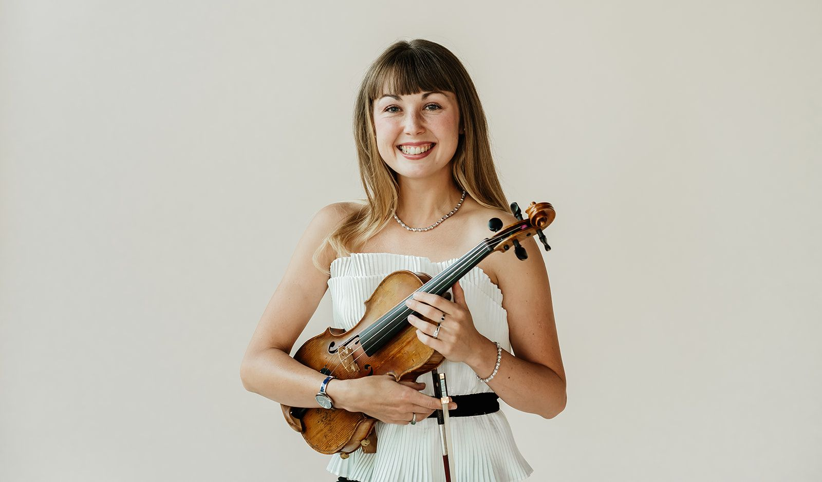 A woman in a white tank top is holding a violin and smiling.