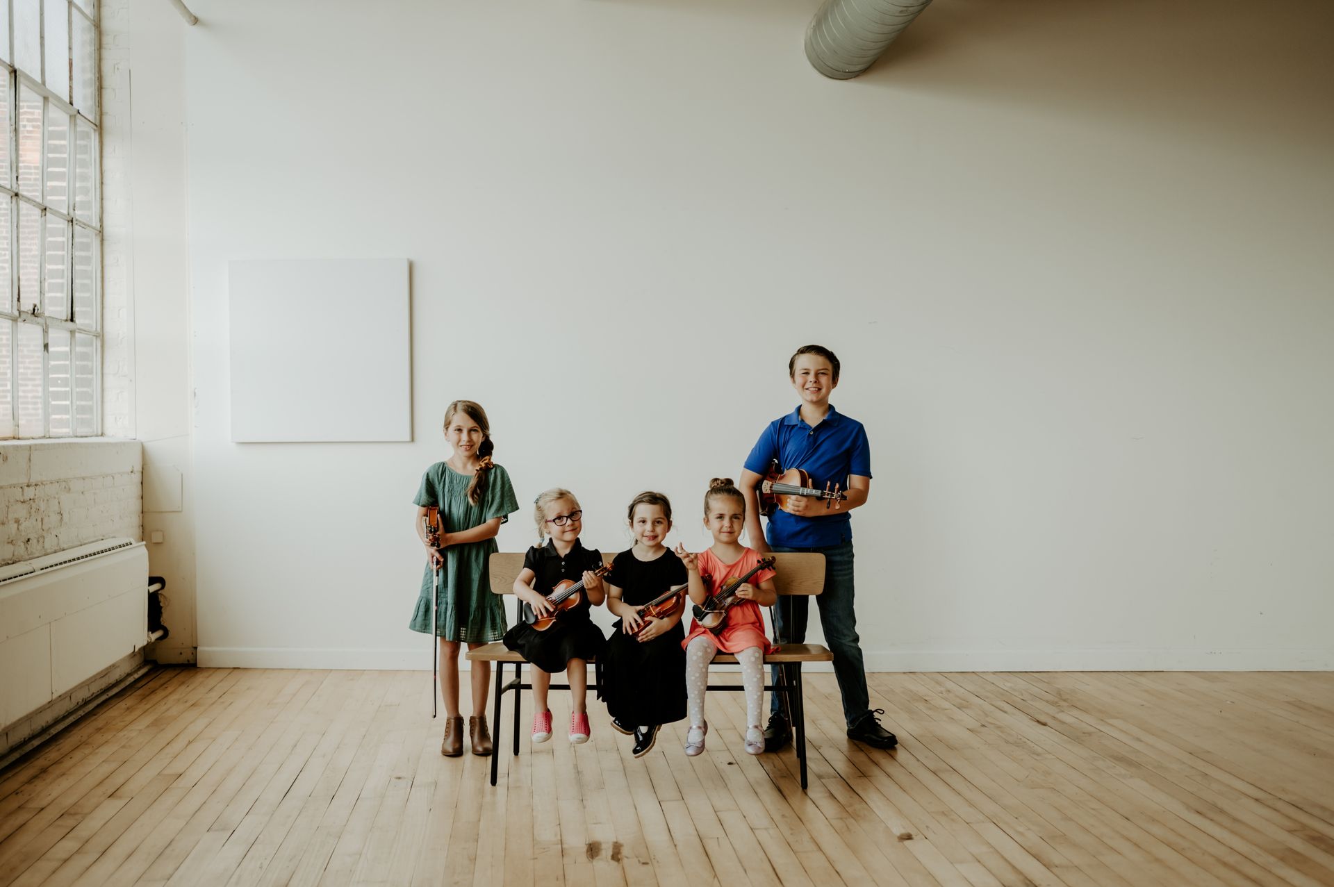 A group of children are sitting on a bench holding violins.