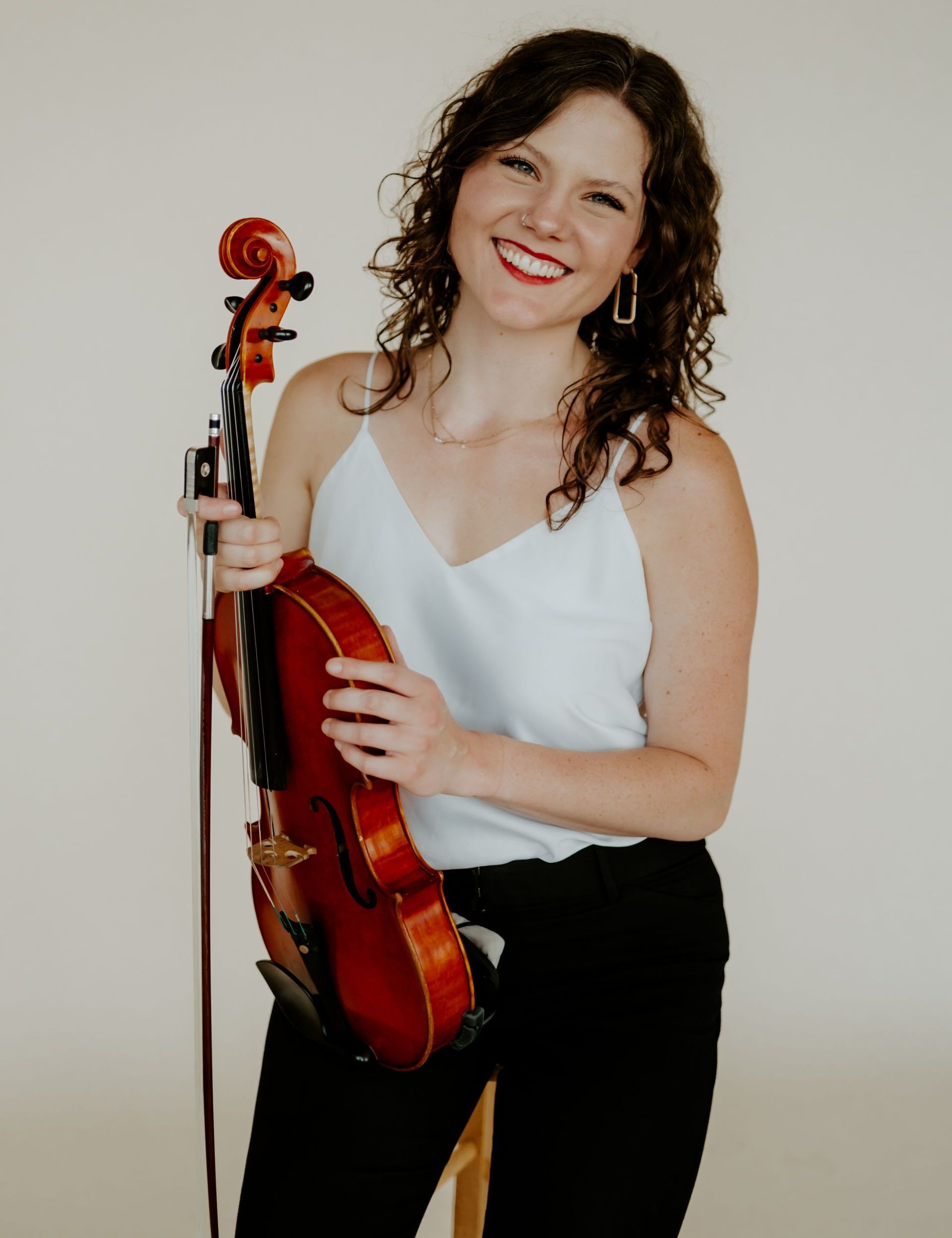 A woman in a white tank top is holding a violin and smiling.