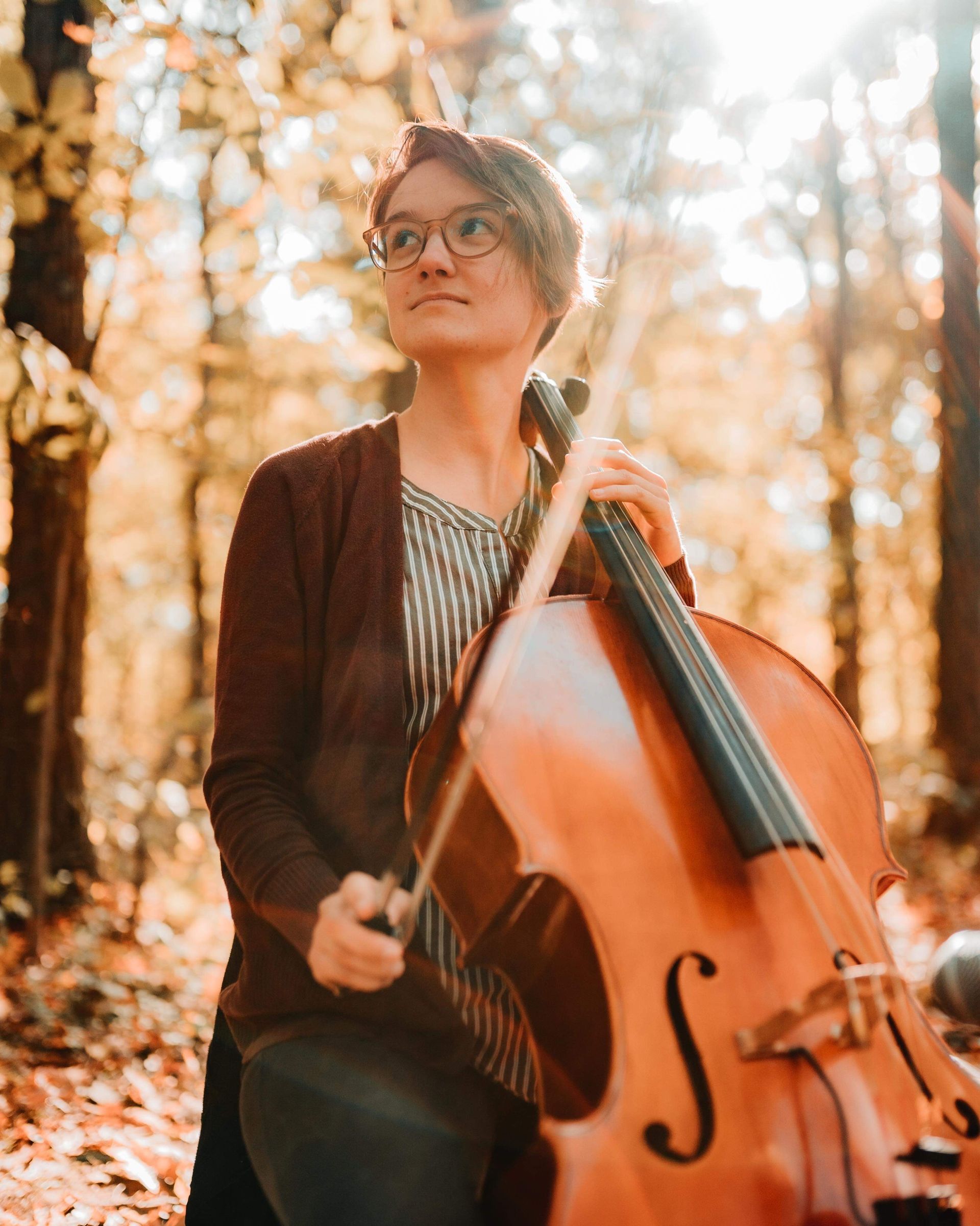 A woman is holding a cello in the woods.