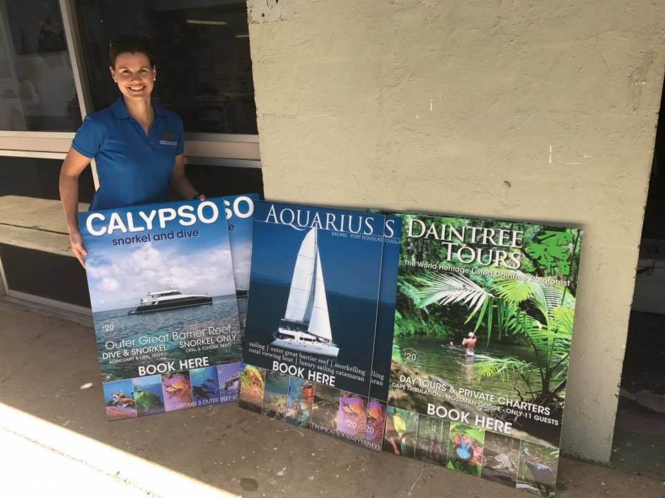 A woman is holding three posters in front of a building- Sponsorship Signage in Port Douglas, QLD