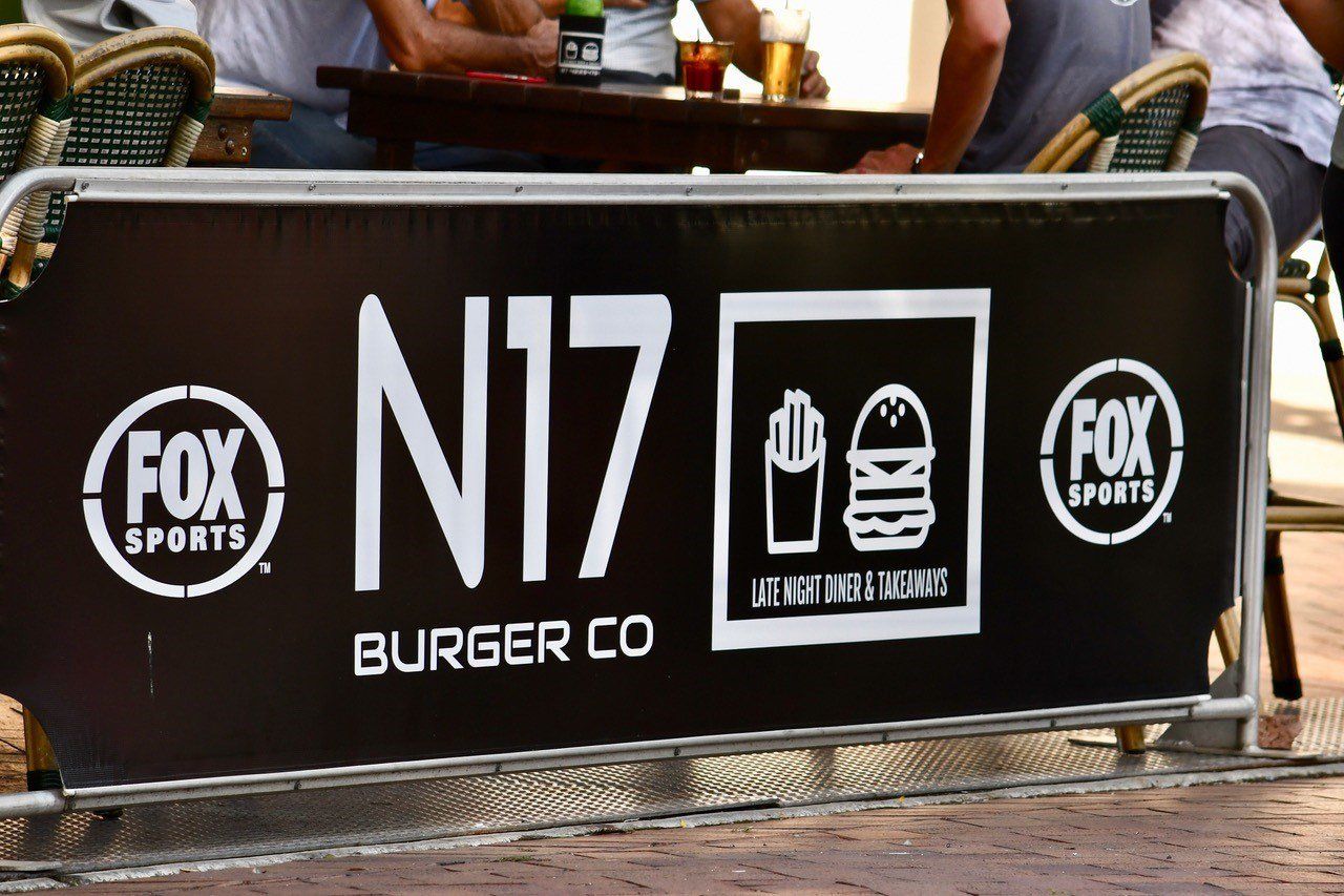 A group of people are sitting at a table in front of a sign for fox sports burger co- Sponsorship Signage in Port Douglas, QLD