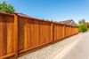 A long, stained wood privacy fence stretches along a gravel border next to a paved road under a clear blue sky.