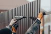 A person wearing gloves uses a power drill to fasten a brown corrugated metal panel to a fence frame.