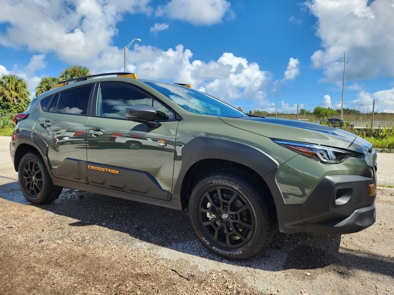 Green SUV with black wheels, roof rack, and side lettering parked on pavement under a cloudy sky.