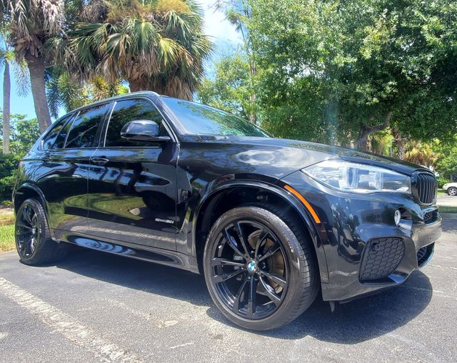 Black BMW X5 SUV parked on a paved surface with trees in the background under a bright sky.