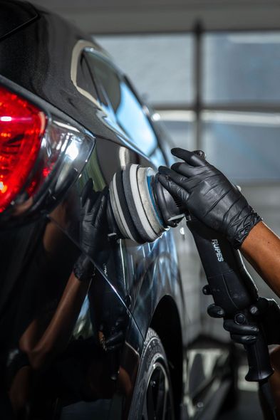 Hands wearing black gloves using an orbital polisher on a black car, detailing the paint.