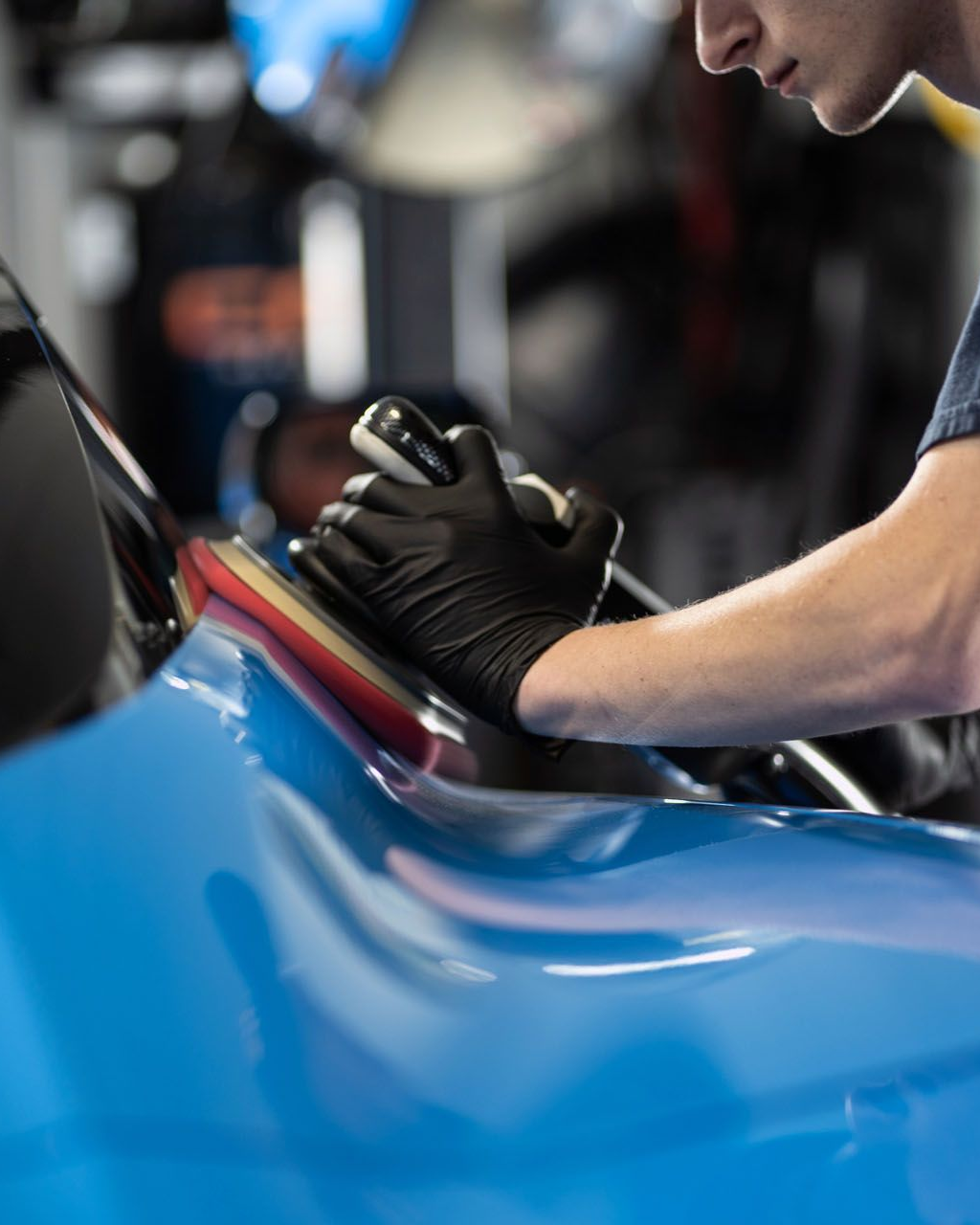 Person polishing a blue car with a handheld buffer in a garage.