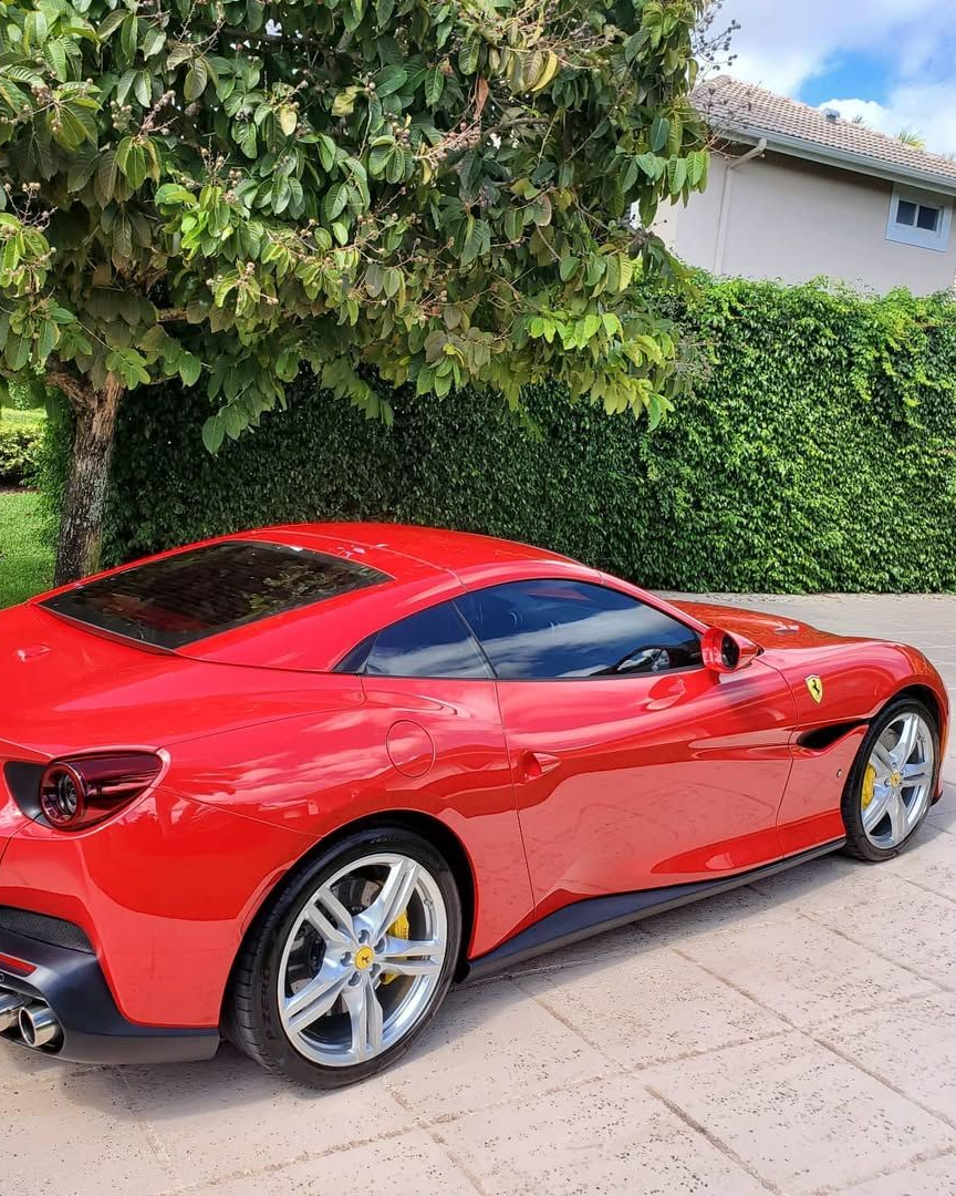 Red Ferrari parked near lush green foliage and a building.