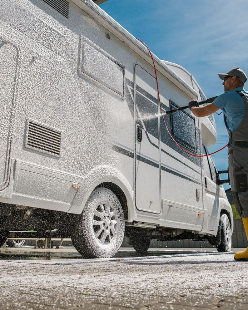 Person washing a white RV with soapy water under a blue sky.