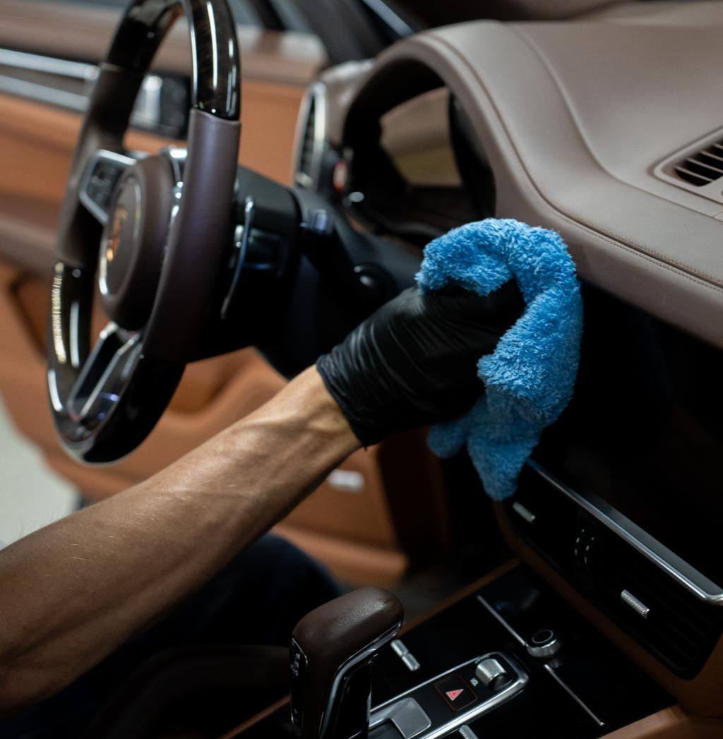Hand wearing black glove wiping car's brown dashboard with blue cloth. Steering wheel and gear shift visible.