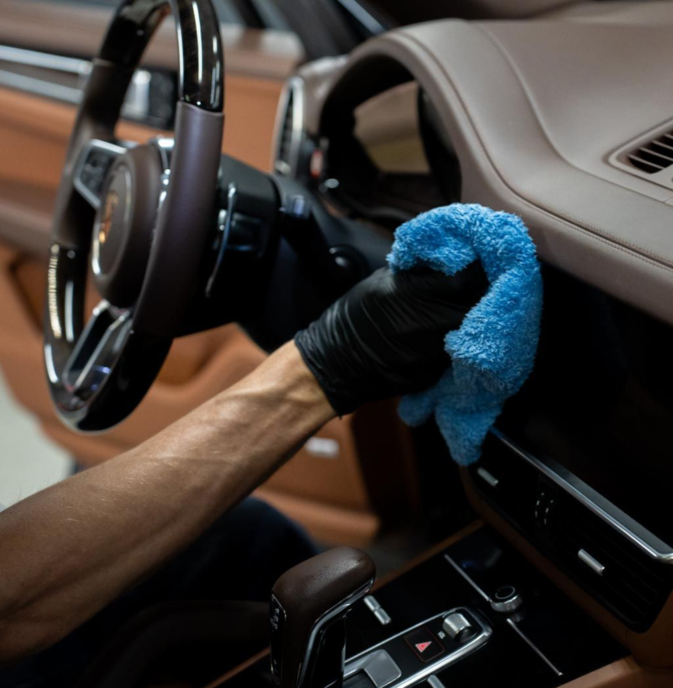 Hand wearing black glove wiping car's brown dashboard with blue cloth. Steering wheel and gear shift visible.