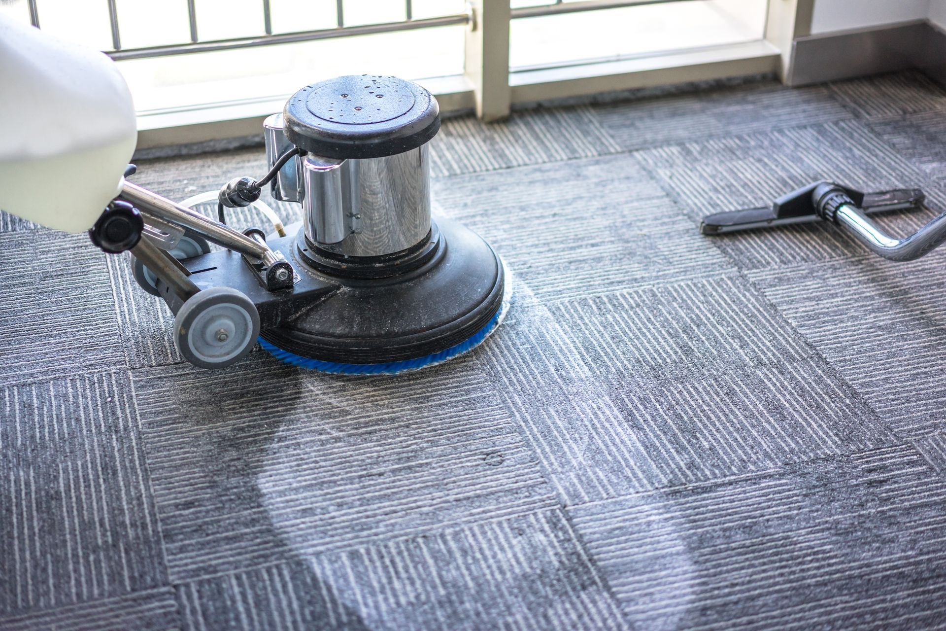 Carpet cleaning machine scrubbing a gray and blue carpeted floor near a window. Carpet cleaning machine scrubbing a gray and blue carpeted floor near a window.