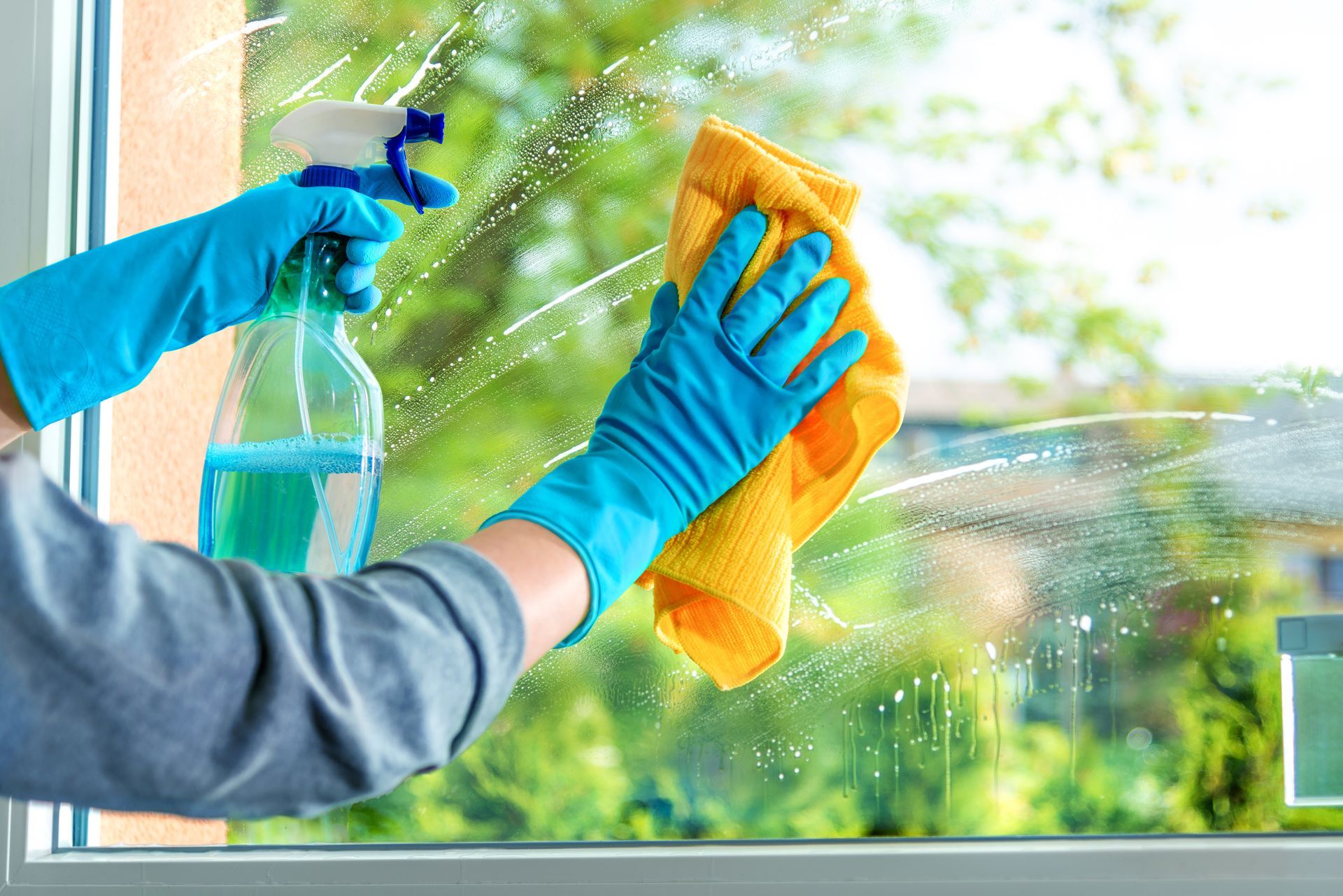 Hands in blue gloves cleaning window with spray bottle and yellow cloth. Hands in blue gloves cleaning window with spray bottle and yellow cloth.