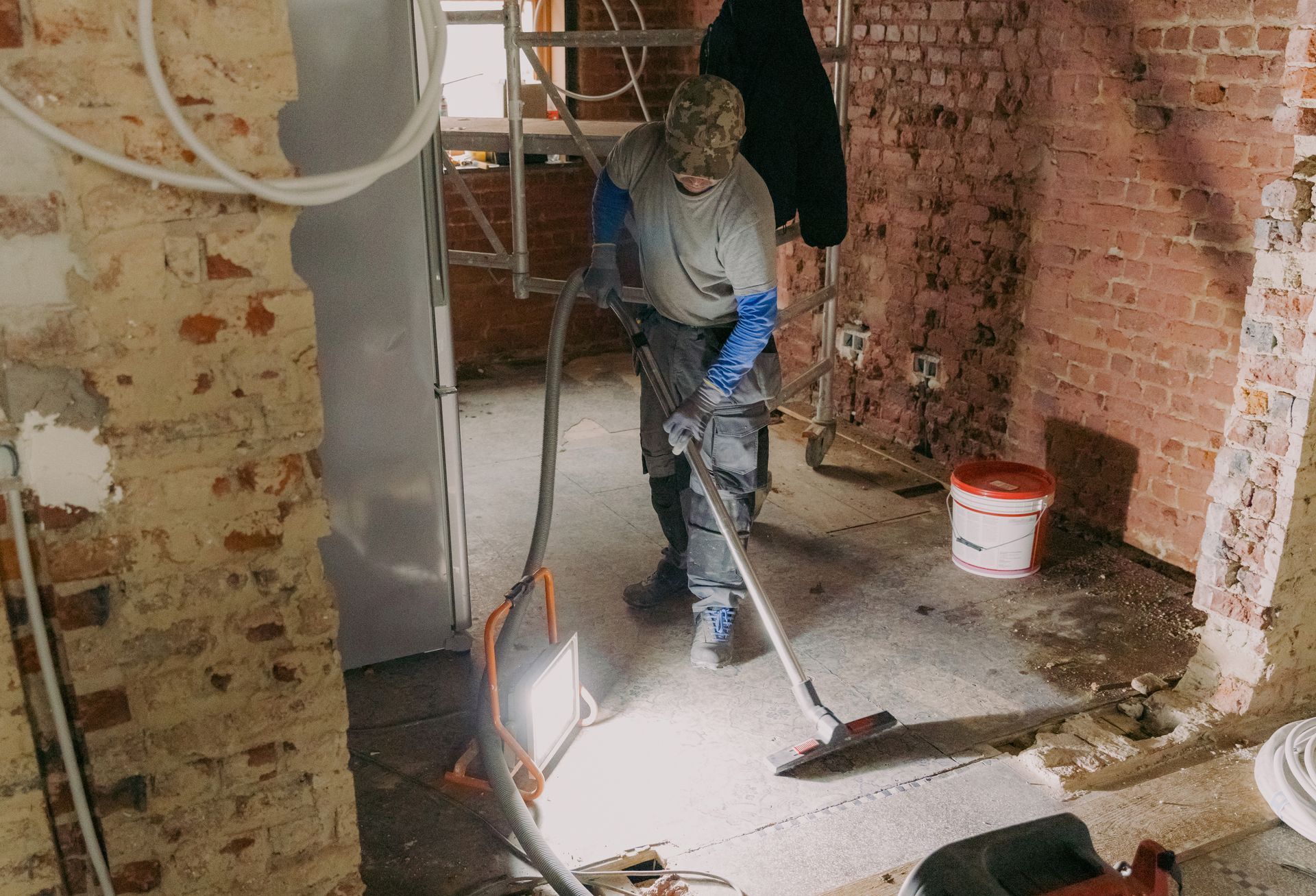 Man in work clothes smoothing floor in a room under construction with exposed brick walls.