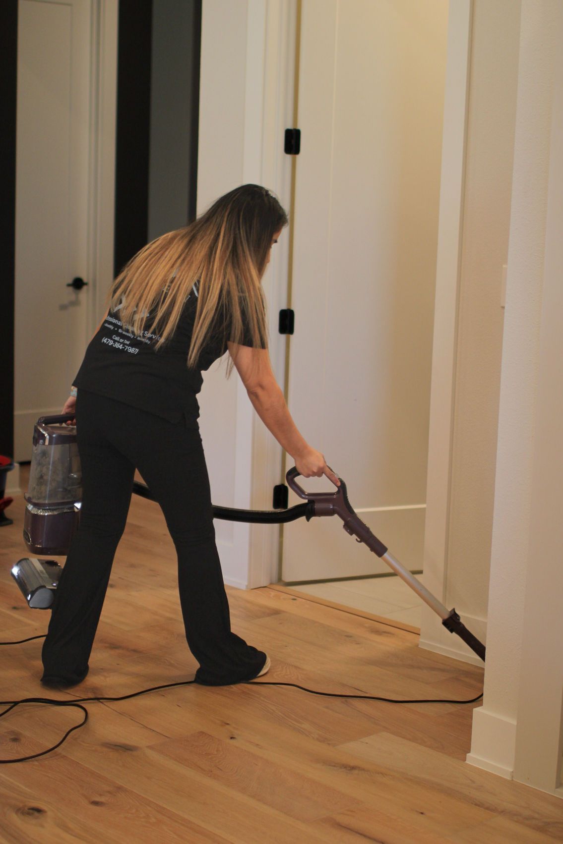 Person vacuuming a wooden floor in a hallway with white walls and a doorway.