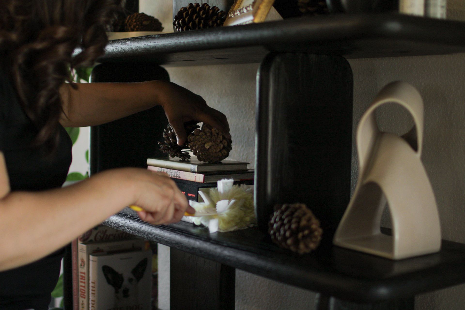 A person dusting a shelf with decor, including books, pinecones, and sculptures.