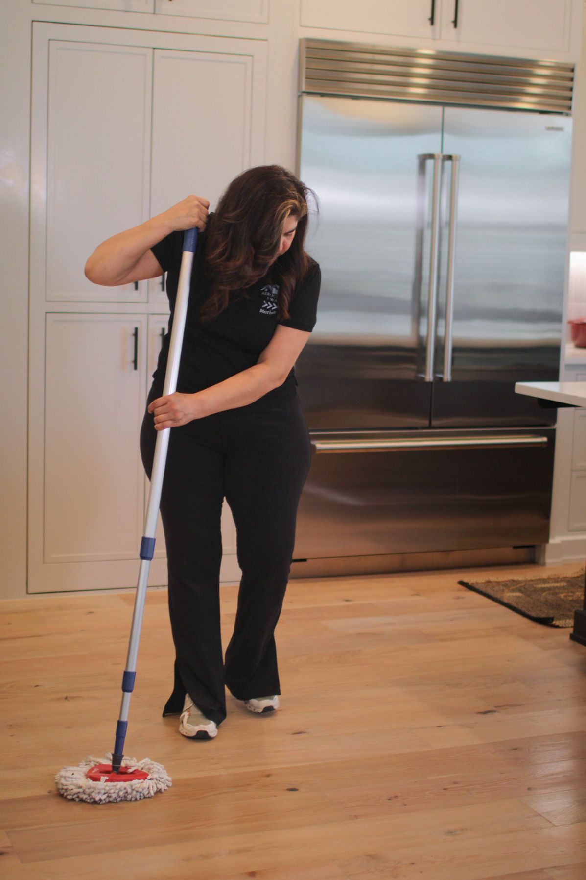 Woman mopping light wood floor in a kitchen with a stainless steel refrigerator and white cabinets.