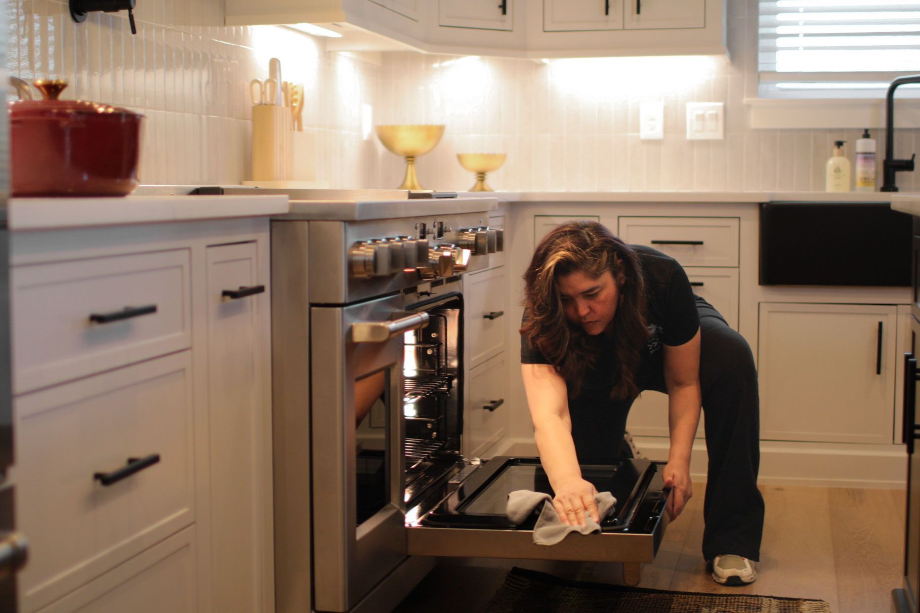 Woman cleaning the inside of a stainless steel oven in a white kitchen with black hardware.