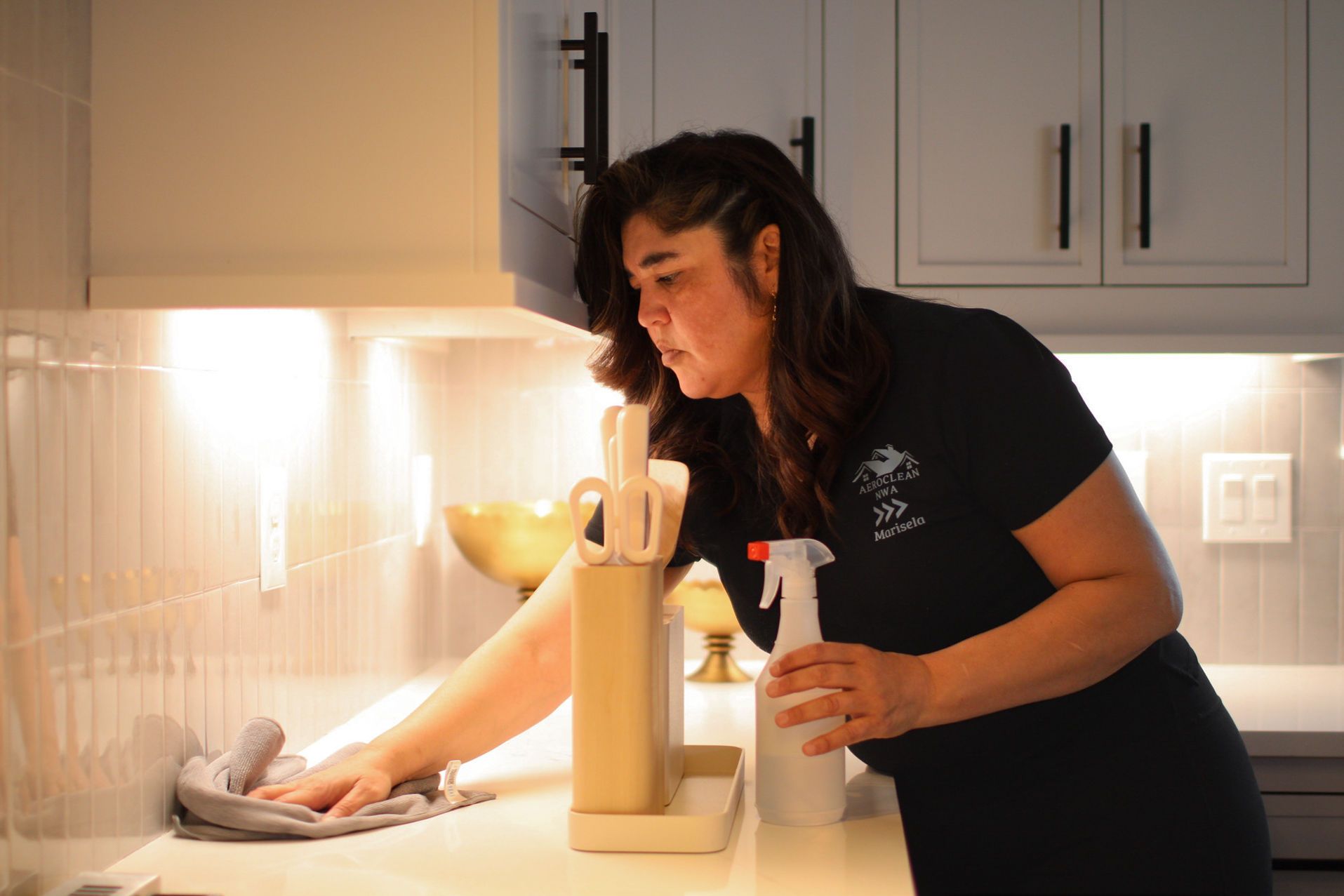 Woman cleaning a kitchen countertop with a spray bottle and cloth, next to a countertop appliance.