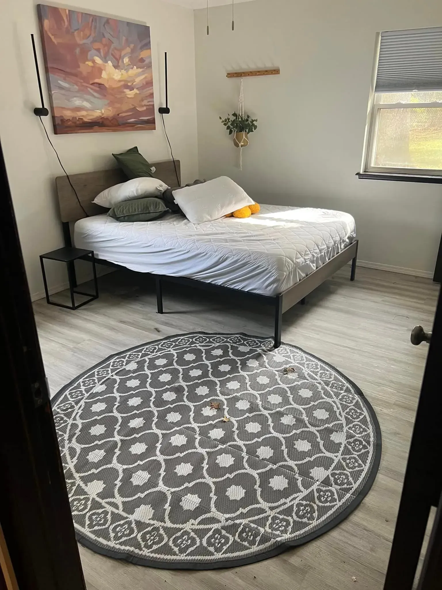 Bedroom with bed, rug, painting, and hanging plant; neutral tones.