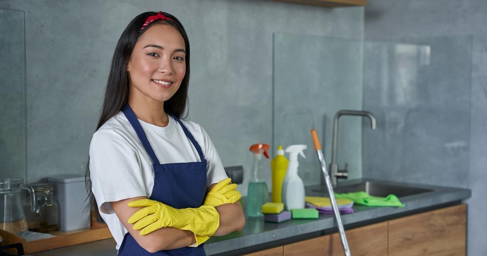 Woman in apron and gloves, smiling, in a kitchen setting with cleaning supplies.