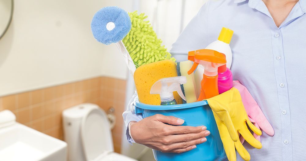 Person holding a blue bucket filled with cleaning supplies in a bathroom setting.