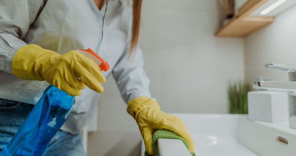Woman in yellow gloves cleaning a white bathroom sink with spray bottle and sponge.