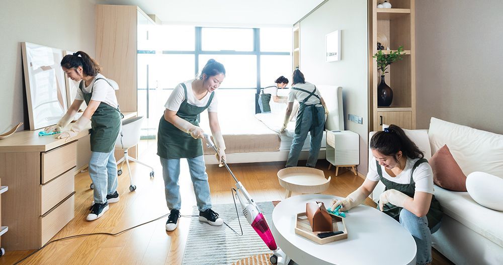 People cleaning an apartment: dusting furniture, vacuuming carpet, and wiping surfaces. Bright interior.