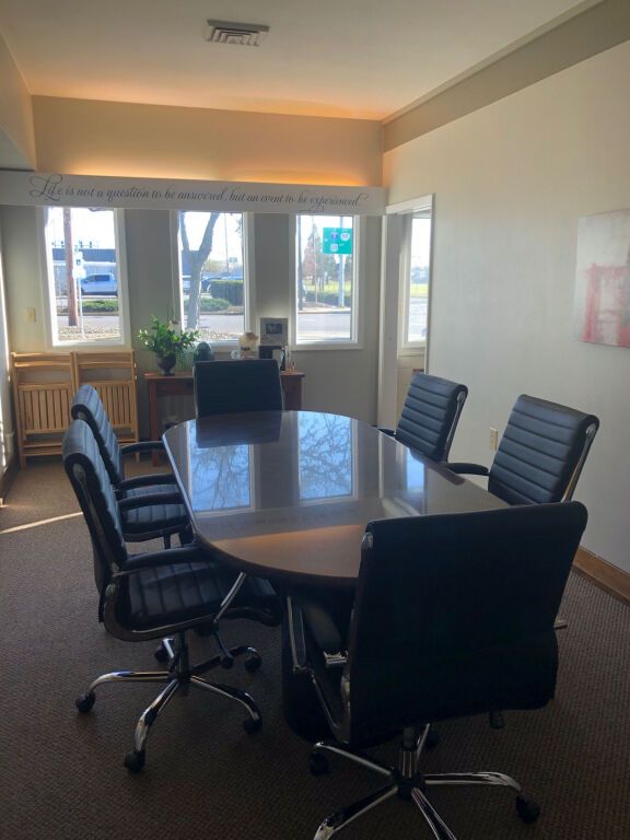 Conference room with oval wooden table, black chairs, and windows with sunlight.