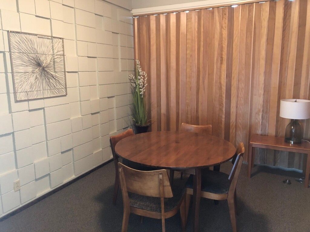 A dining room with a round wooden table, chairs, and window blinds. Artwork and a lamp are present.