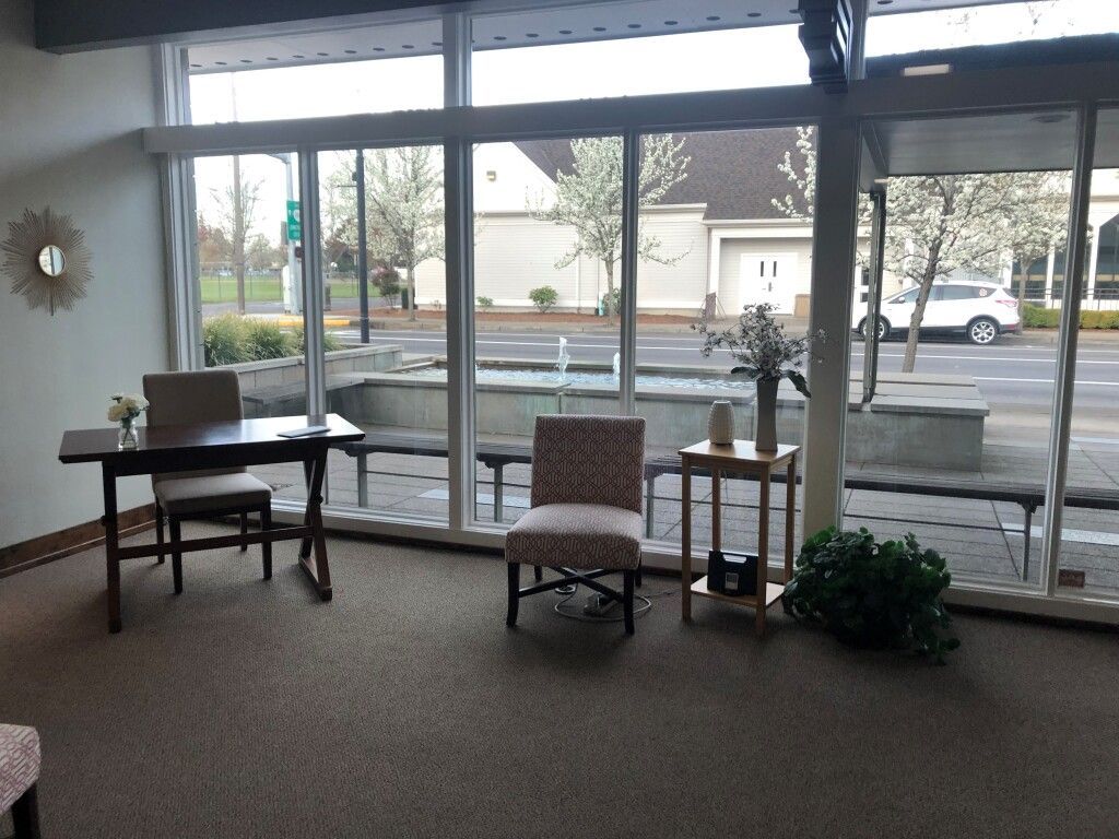 Interior with tables and chairs near large windows overlooking a street with a car and trees.