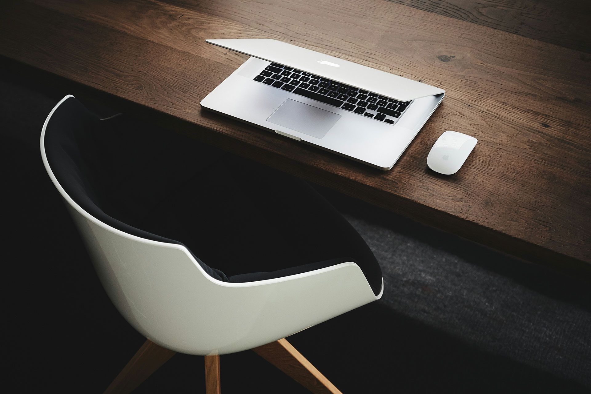 Laptop, white mouse on dark wood desk; white and black chair in the foreground.