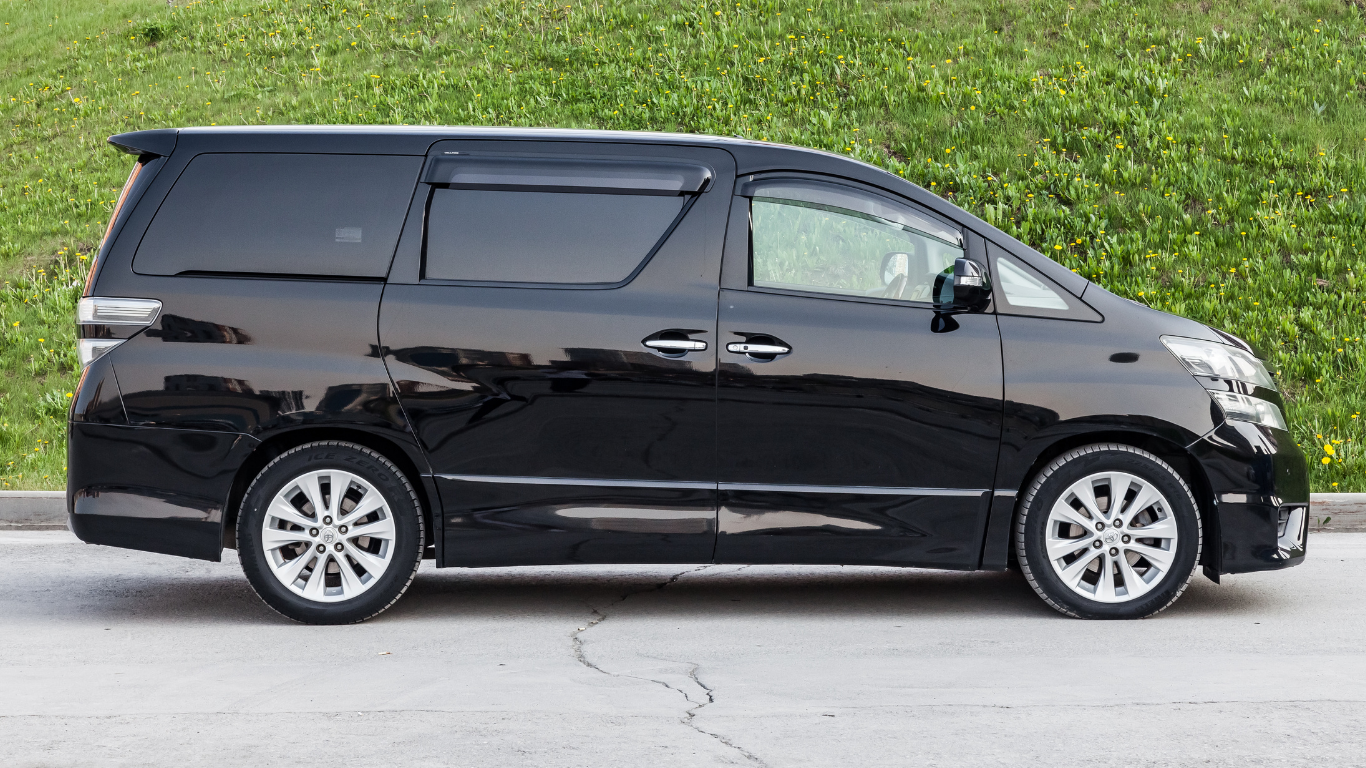 Black minivan parked on a paved road, green grass background.