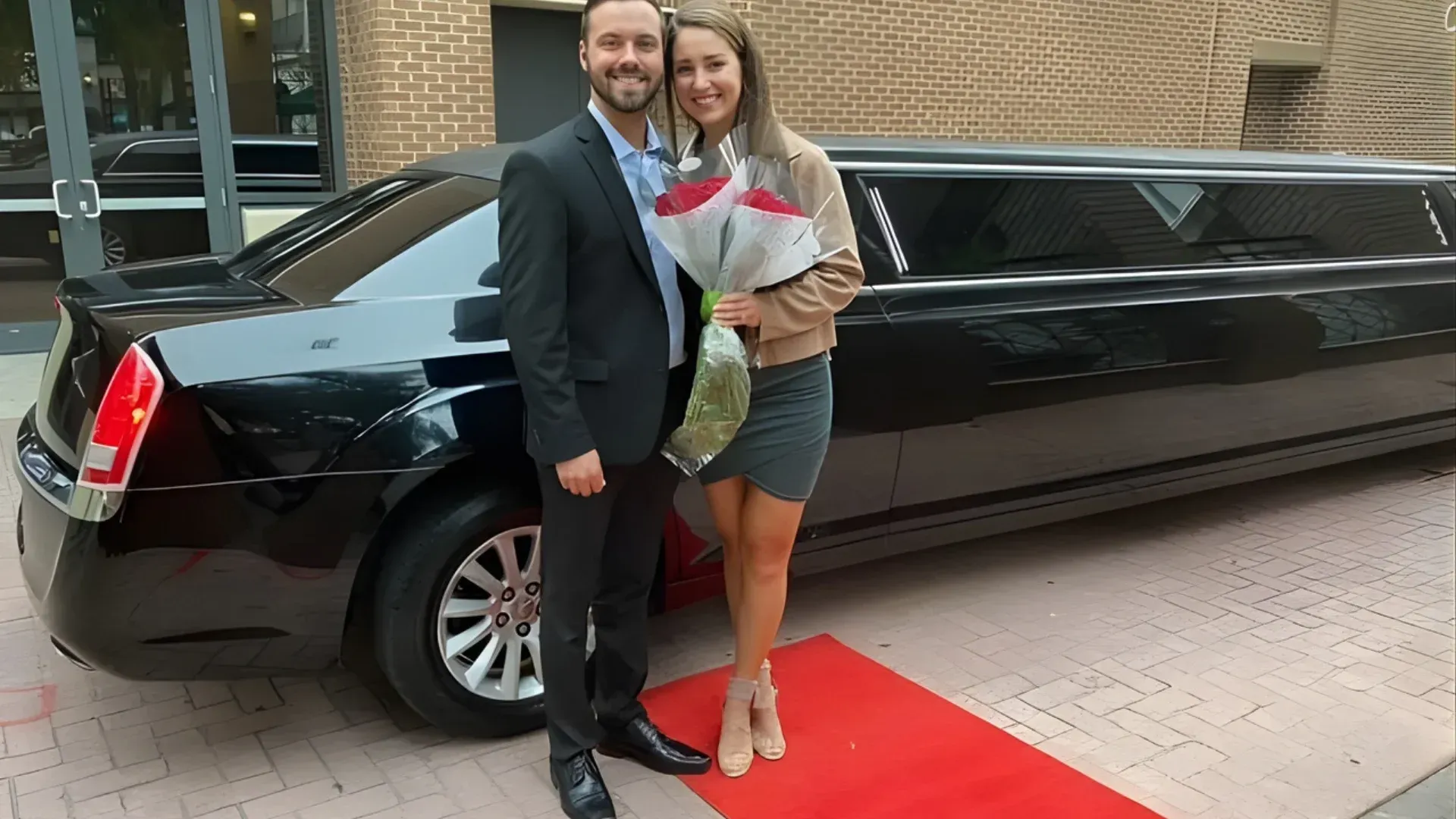 Couple poses by black limousine holding roses on red carpet.
