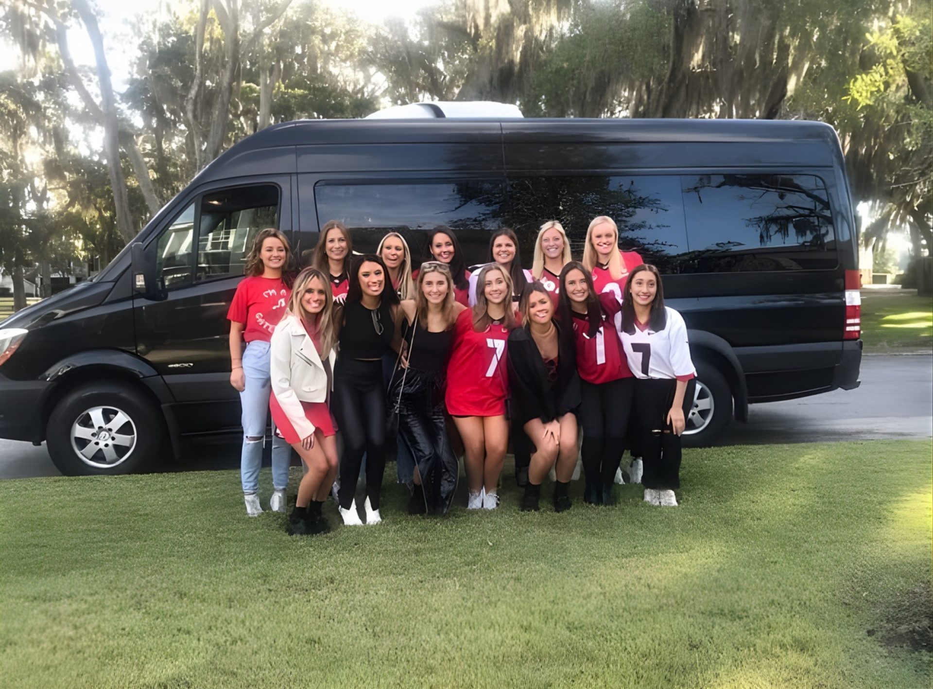 Group of women in front of a black van, posing on green grass outdoors.