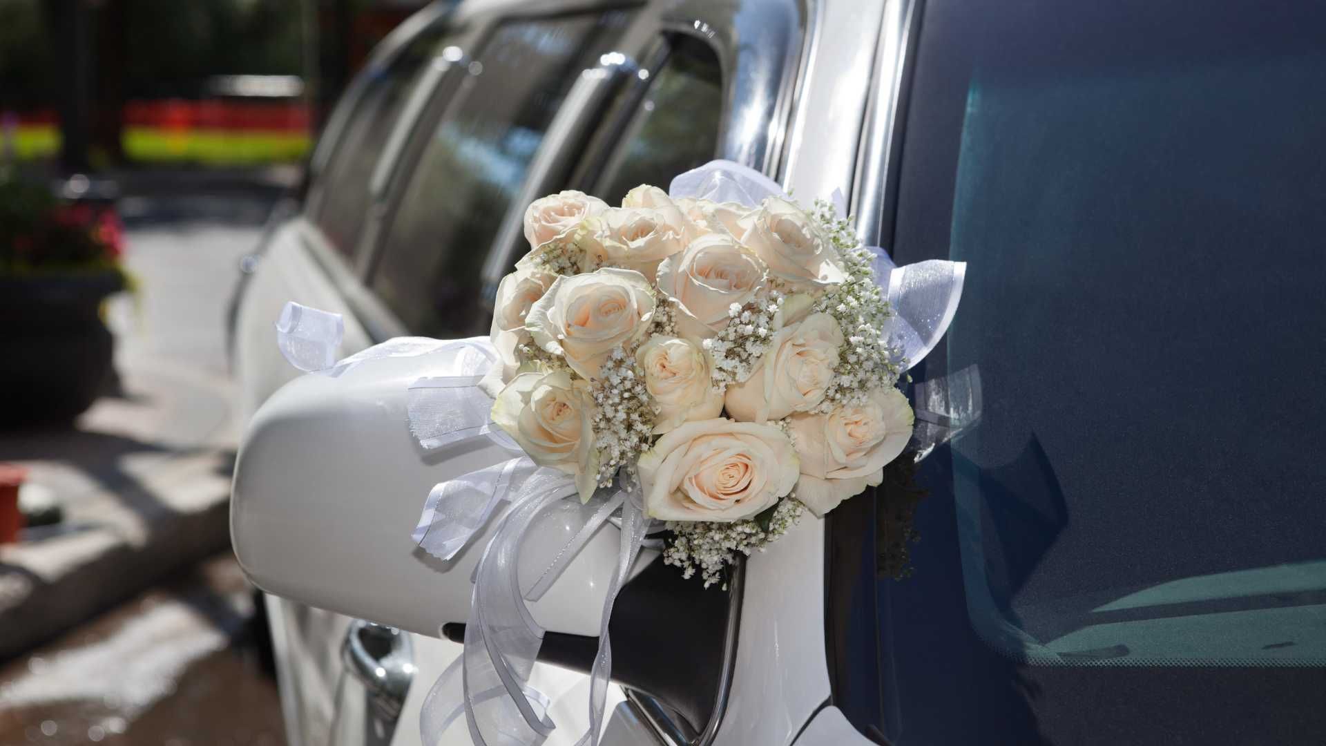 White roses and ribbons decorate a car's side mirror, suggesting a wedding.