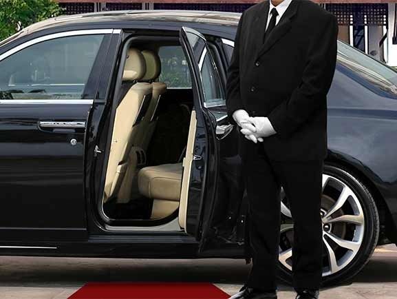 A chauffeur in black suit and white gloves stands by a black car with an open door and red carpet.