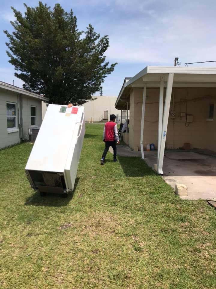 A man is carrying a refrigerator in a backyard.