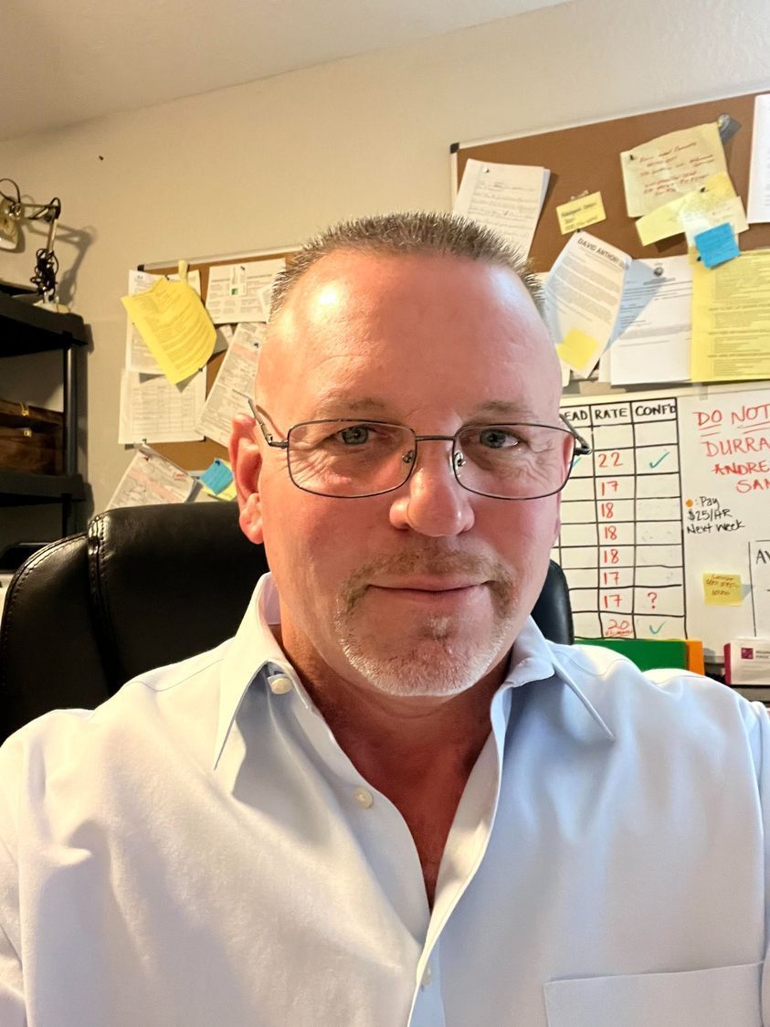 A man wearing glasses is sitting in front of a bulletin board with sticky notes on it