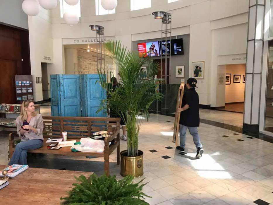 A woman is sitting on a bench in a lobby while a man moves a plant. 28.4845° N, 81.2519° W