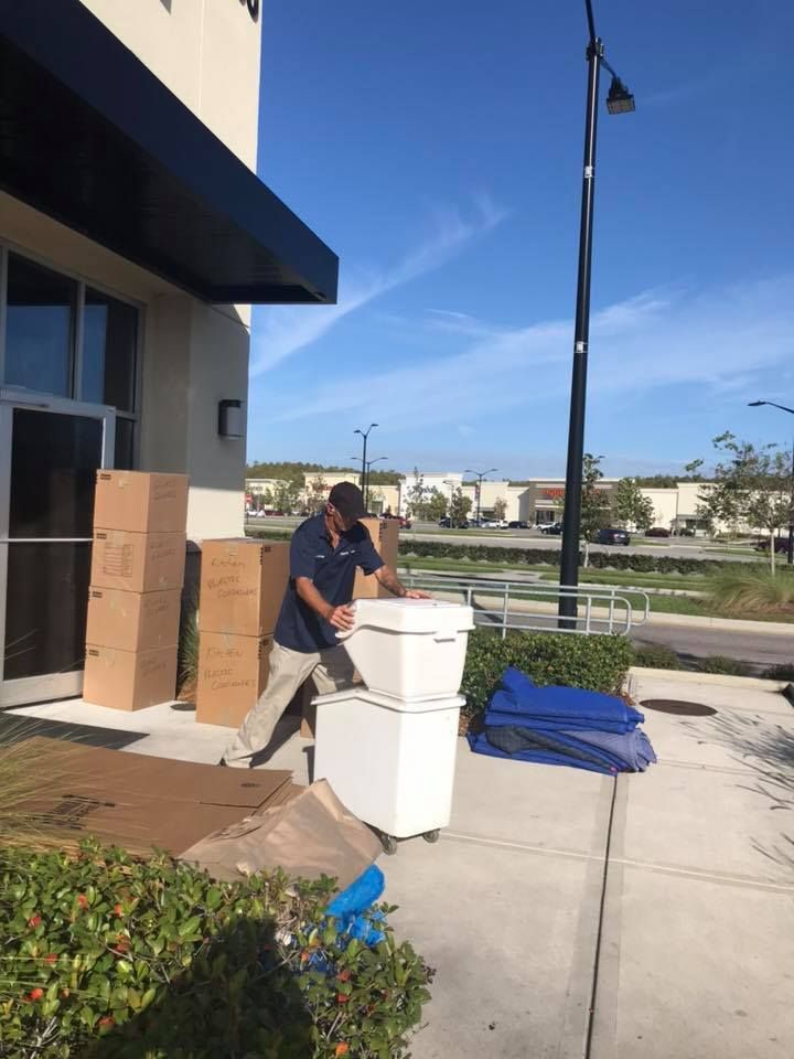 A man is loading boxes into a white bin on a sidewalk in front of a building. 28.4845° N, 81.2519° W