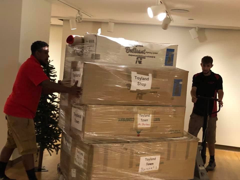 Two men are moving a stack of boxes in a room. 28.4845° N, 81.2519° W