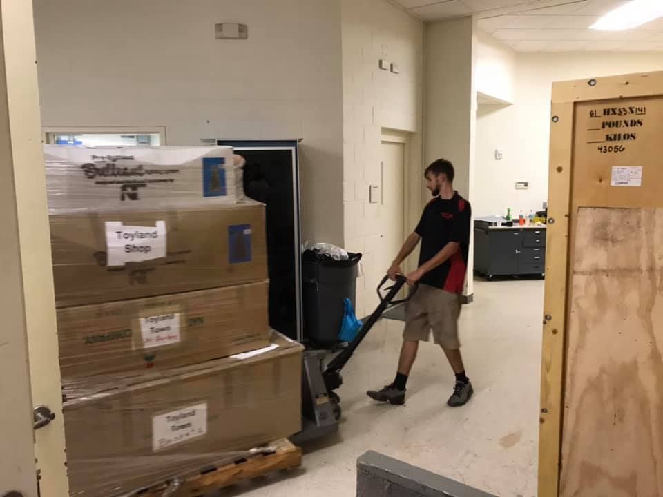 A man is pushing a pallet truck with boxes on it in a room. 28.4845° N, 81.2519° W