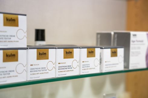 Eine Reihe weisser Kulm-Hautpflegeproduktverpackungen mit goldenen Etiketten ist auf einem Glasregal angeordnet.