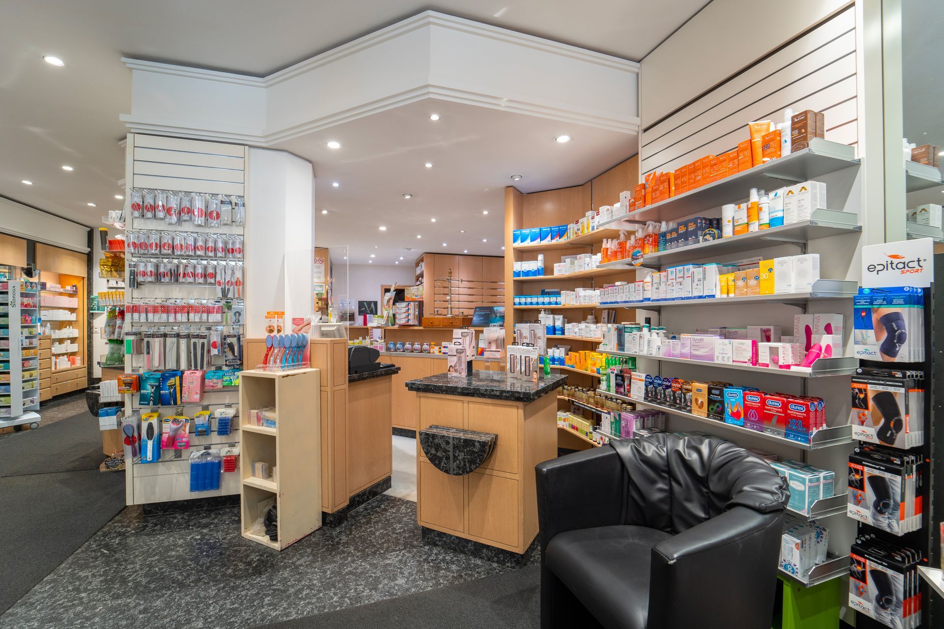 Das helle Interieur einer Apotheke mit Produktregalen, einem Kundendienstschalter und einem schwarzen Sessel im Vordergrund.