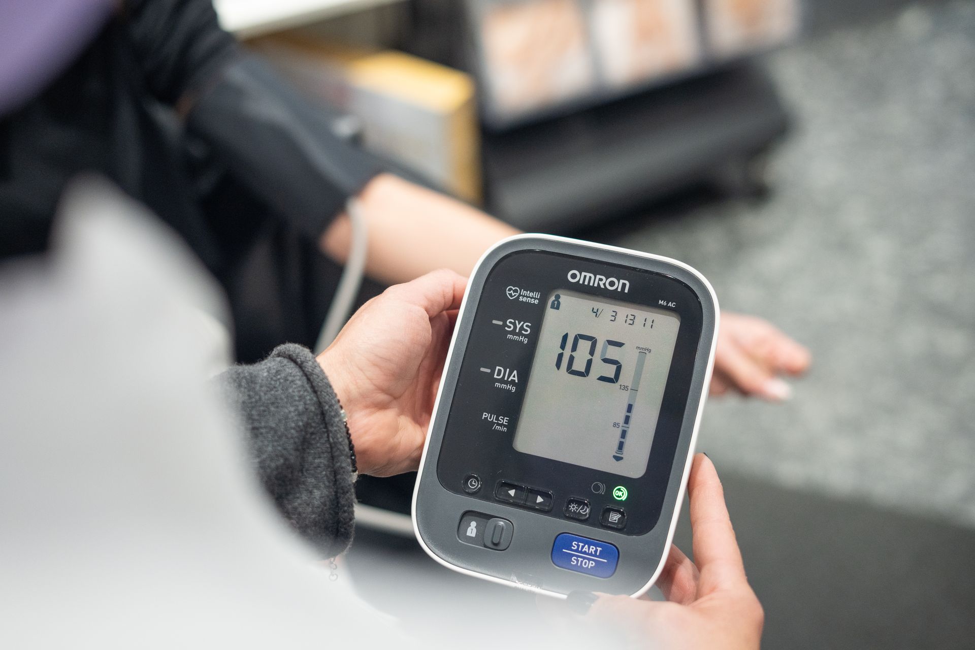 Person hält Omron-Blutdruckmessgerät mit Anzeige 105 mmHg, andere Person trägt Blutdruckmanschette, medizinische Messung.