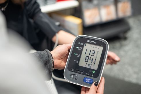 Eine Person hält ein digitales Blutdruckmessgerät, das einen Wert von 113/84 mmHg bei einem Puls von 110 anzeigt.