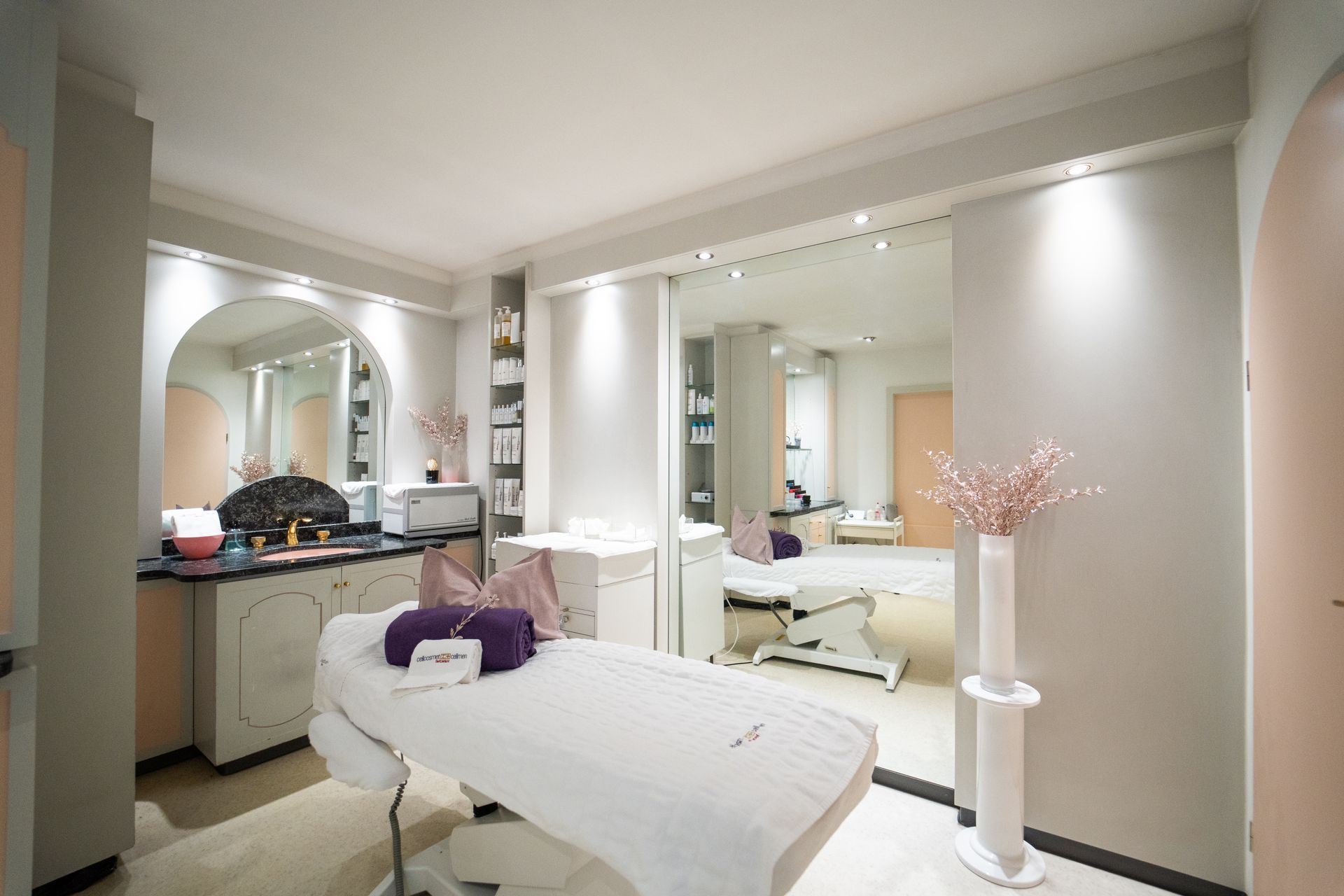 Ein moderner, in neutralen Tönen gehaltener Spa-Behandlungsraum mit Massageliege, Waschbecken und einer grossen Spiegelwand.